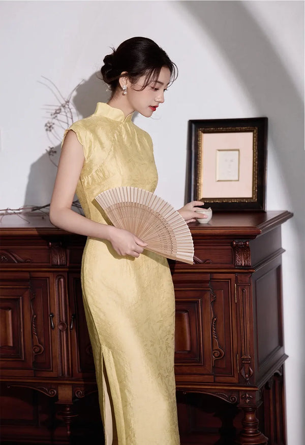 Woman in a Yellow Floral Sleeveless Midi Cheongsam Qipao Dressholding a fan in a room with wooden furniture and a framed picture.