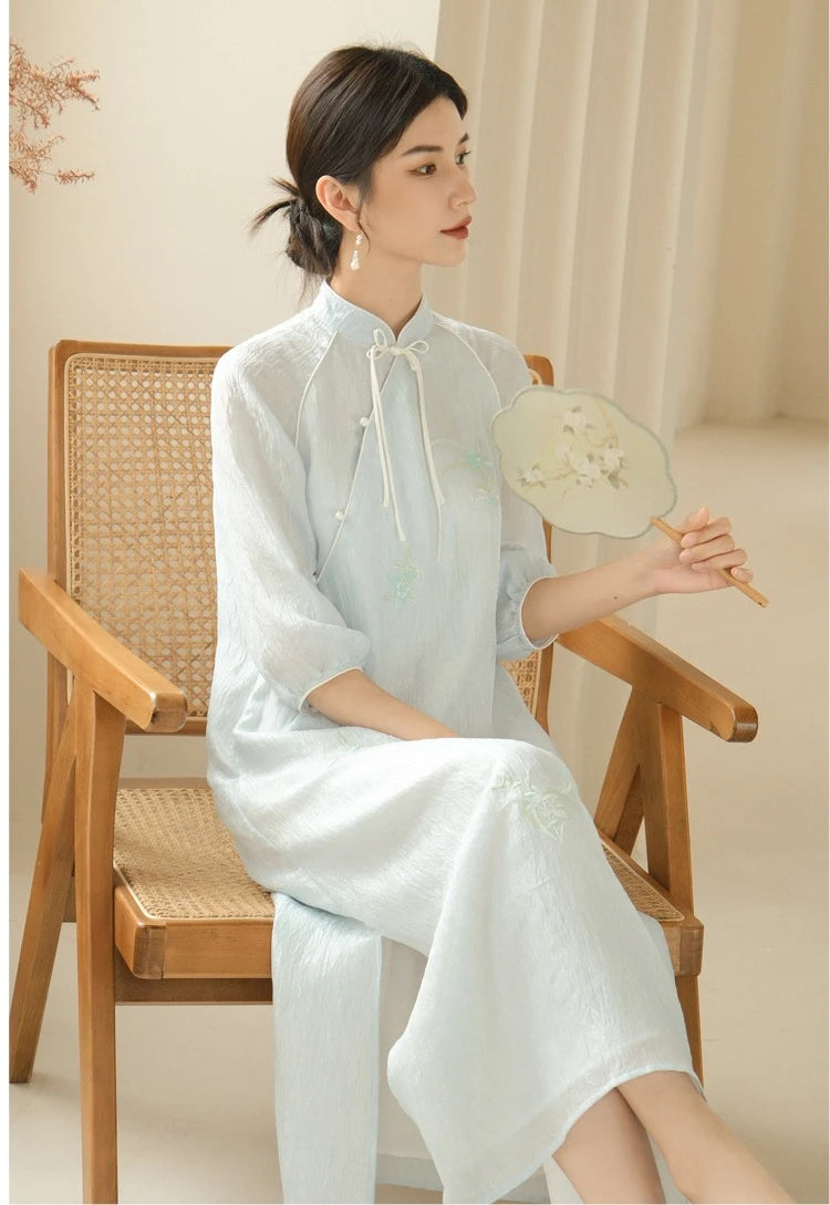 a chinese Woman in a Zen-Inspired Light Blue Loose Cheongsam Qipao Dress  sitting on a wooden chair holding a fan.
