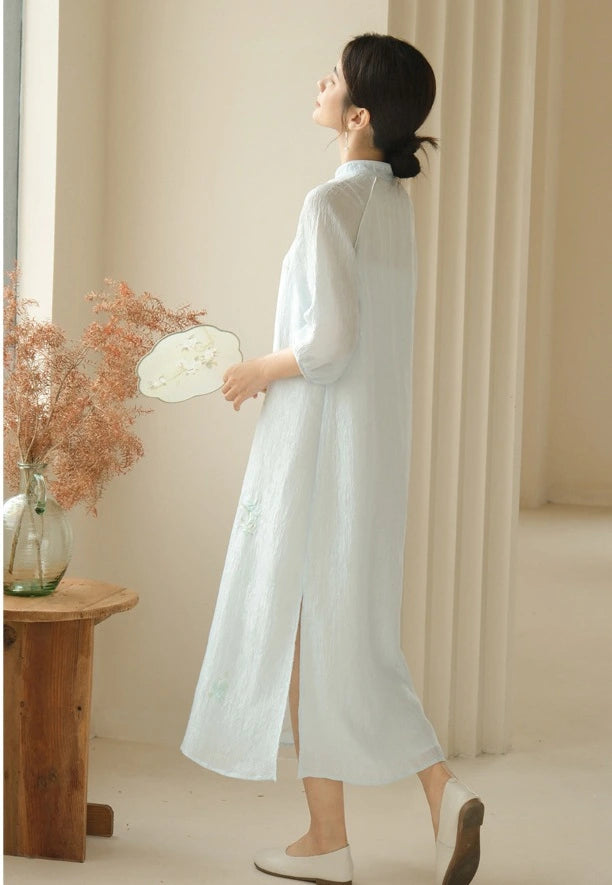 Woman in a long Zen-Inspired Light Blue Loose Cheongsam Qipao Dress  standing in a softly lit room with a wooden table and decorative plants.