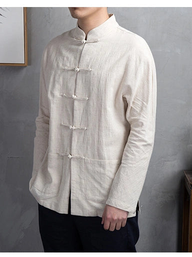 A man wearing a light beige, long-sleeved Chinese-style jacket with traditional frog closures and a mandarin collar, paired with dark pants, standing against a textured gray wall.
