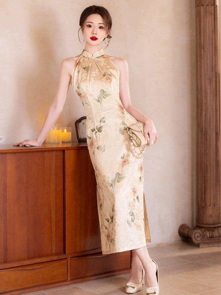 A woman models a sleeveless beige cheongsam dress with a green and pink floral pattern, standing in a warm-toned indoor setting next to a wooden cabinet.