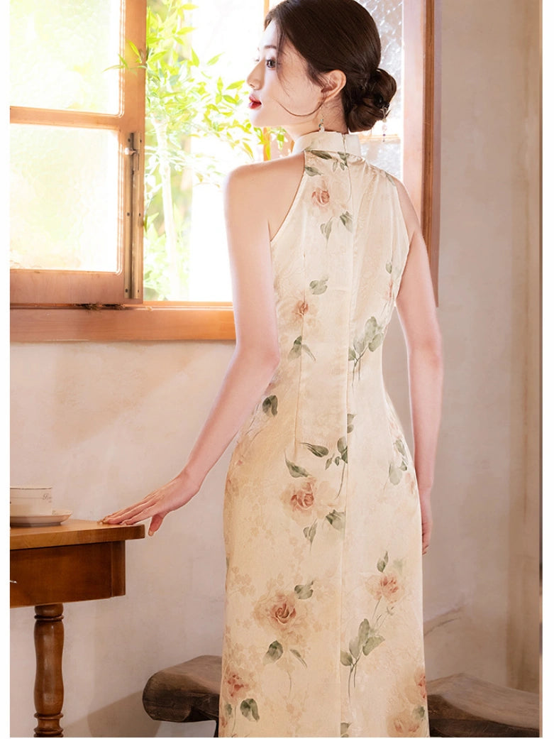 A woman wearing a sleeveless cream-colored cheongsam dress with a delicate pink and green floral print, standing in a sunlit room next to a wooden table.