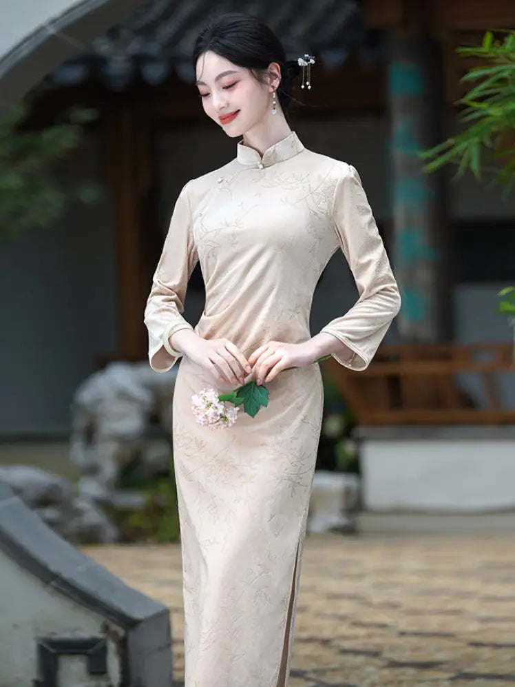 A woman wearing a beige long-sleeve  Velvet  Qipao cheongsam dress with subtle floral embroidery, standing in a traditional courtyard setting.
