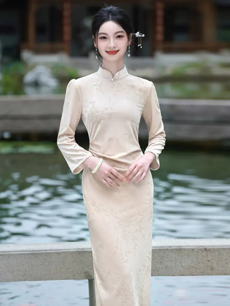 A woman wearing a beige long-sleeve Velvet Long Sleeve Qipao  dress with a high collar, standing outdoors near a pond with a blurred traditional building in the background.