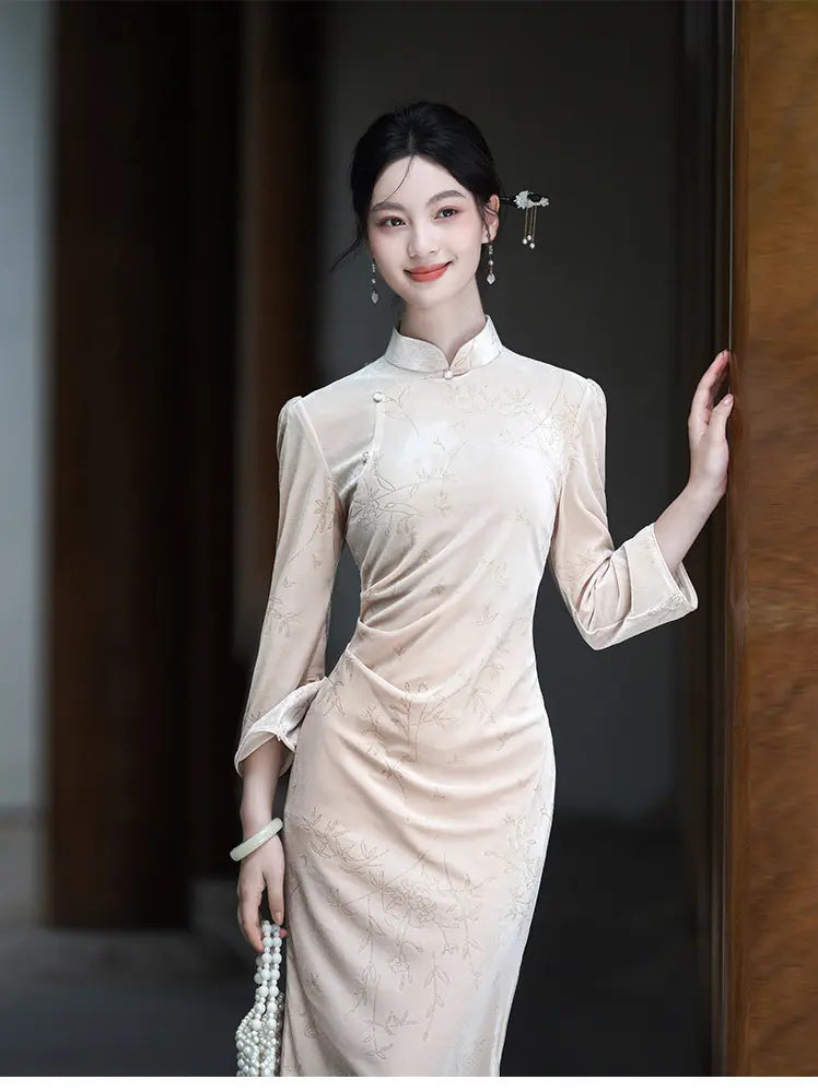 A woman wearing a light beige, long-sleeved Chinese Velvet Qipao cheongsam dress with subtle floral embroidery, posing in a dimly lit corridor.