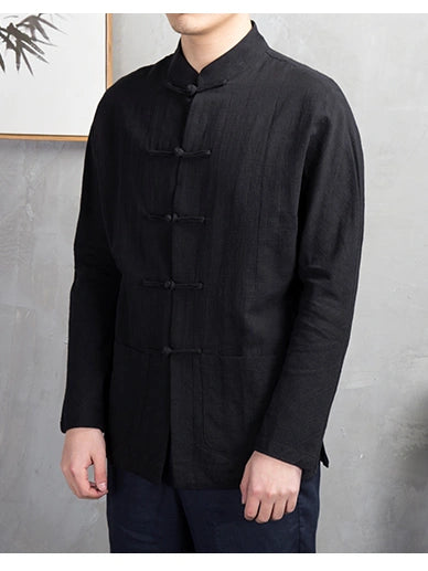 A man wearing a black long-sleeve traditional Chinese-style shirt with a mandarin collar and dark wooden toggle closures, paired with dark pants, standing against a textured gray wall.