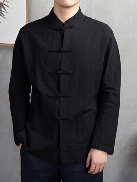 A man wearing a black long-sleeve mandarin collar shirt with traditional Chinese frog closures, paired with dark trousers, standing against a textured gray wall.