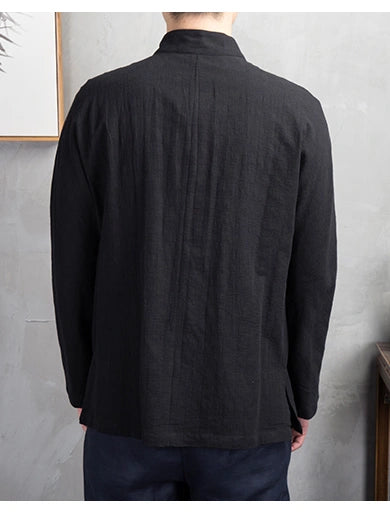 A man viewed from the back wearing a black long-sleeve linen shirt with a mandarin collar and vertical pleats, paired with dark trousers, set against a textured gray wall.