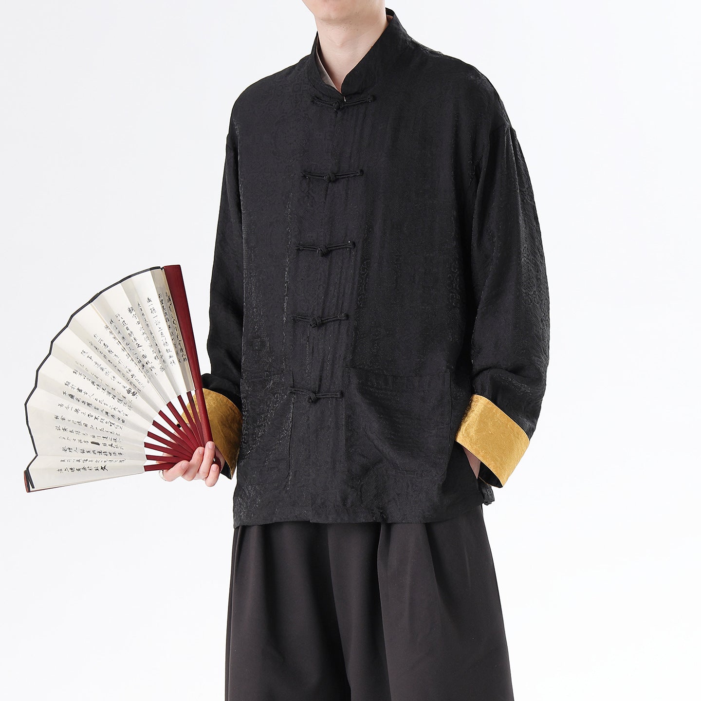 Person wearing a black traditional  Men's Oriental Jacquard Kung Fu Tang Shirt outfit with gold cuffs holding a fan on a white background