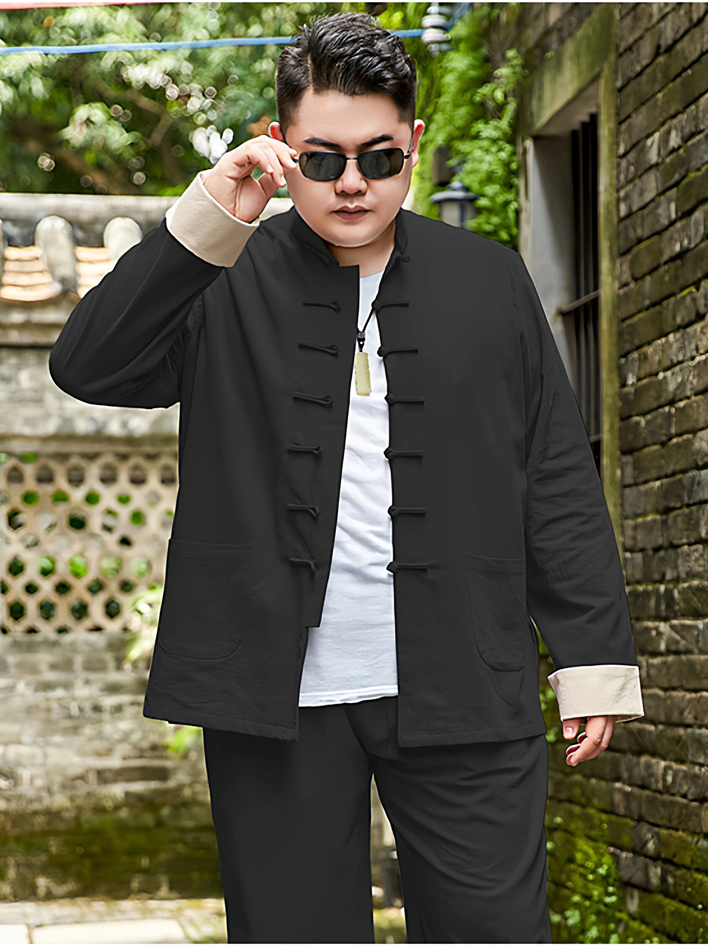 A man wearing a black plus size Chinese-style jacket with beige turned-back cuffs and a white T-shirt underneath, standing outdoors against a brick wall and greenery.