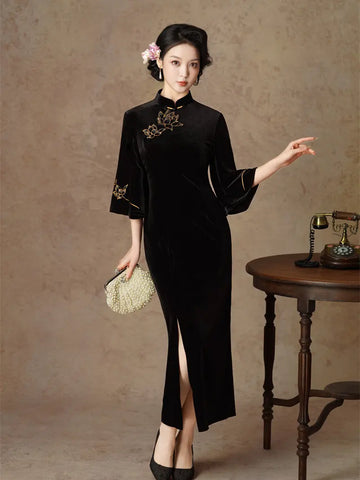 A woman models a black velvet cheongsam-style dress with gold floral embroidery on the chest and sleeve, standing in a studio with a textured beige backdrop, a small wooden table, and a vintage telephone.
