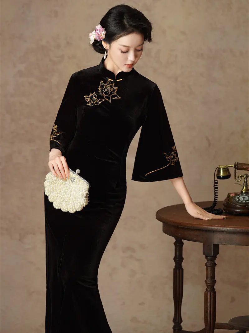 A woman wearing a black velvet qipao cheongsam dress with gold floral embroidery, holding a white textured clutch purse, posed next to a vintage wooden table with a rotary telephone against a textured beige wall.