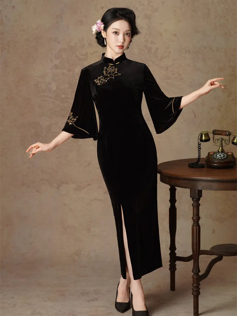 A woman models a black velvet qipao cheongsam-style dress with gold floral embroidery on the chest and sleeve, posed next to a vintage wooden table with a rotary phone against a textured beige backdrop.
