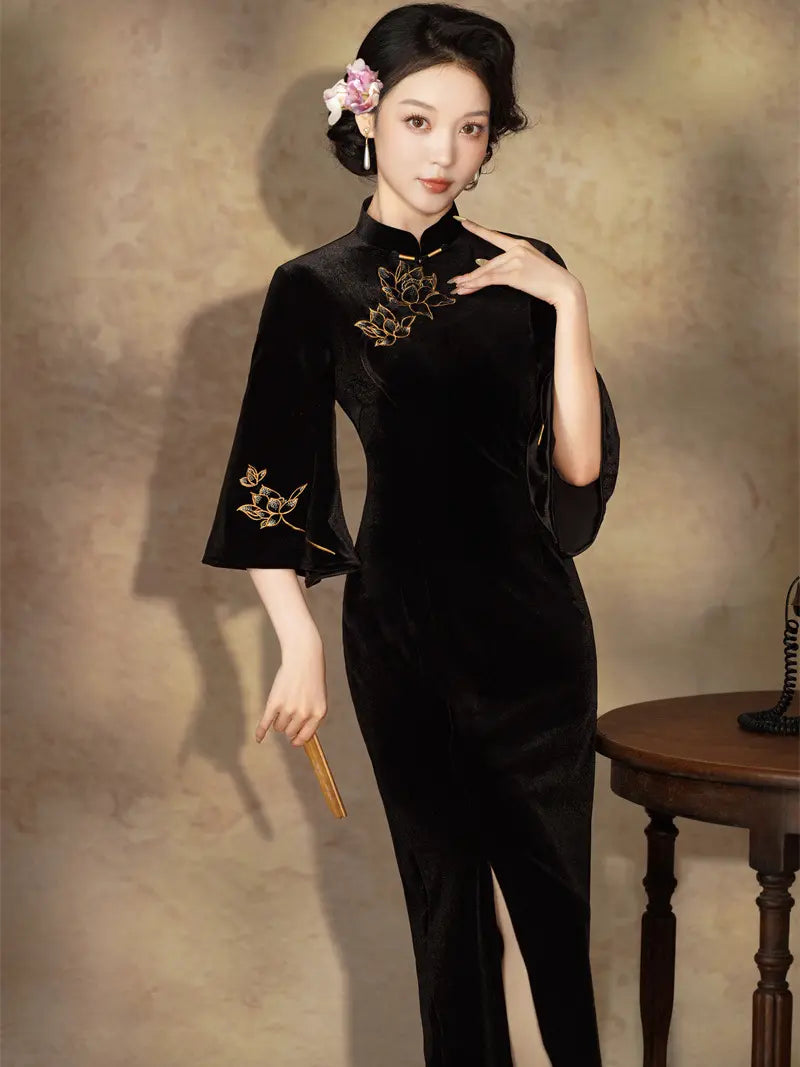 A woman models a black velvet cheongsam with gold floral embroidery on the chest and sleeve, styled with a pink flower in her hair against a textured beige studio backdrop.