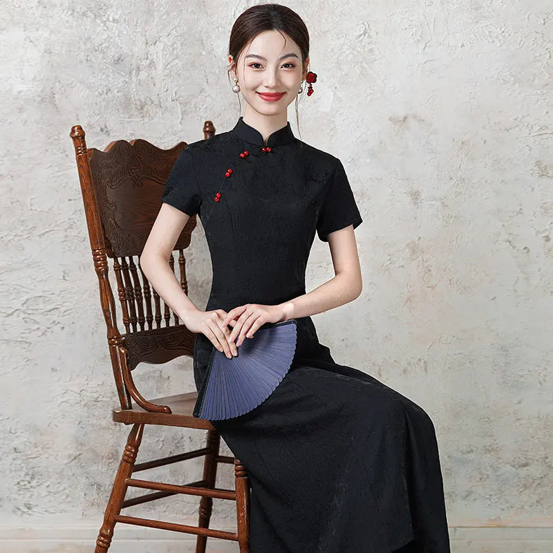 Woman in a black traditional   Short Sleeve Floral A Line Qipao cheongsam outfit  with red accents sitting on a wooden chair against a textured wall.