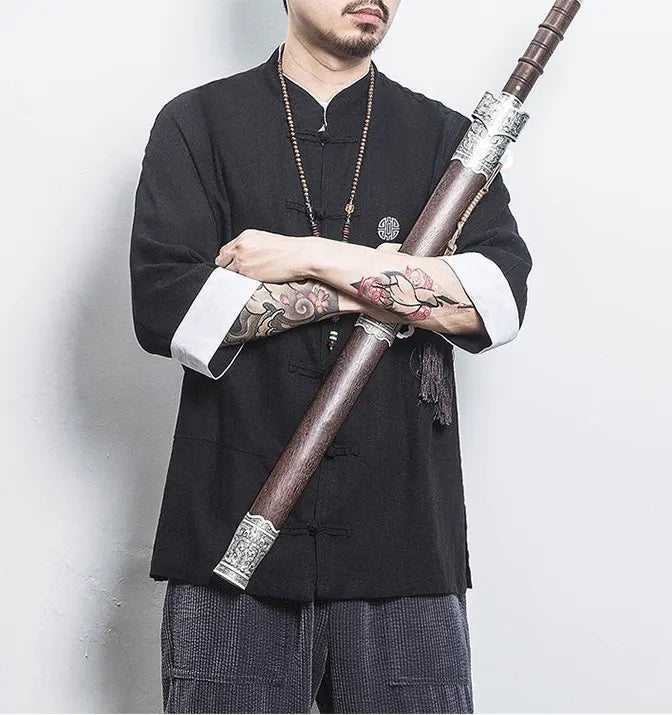 Person wearing a black traditional Three-Quarter Sleeve Zen Heritage Linen-Blend Kong Fu Tang Shirt outfit holding a long, ornate object against a plain background