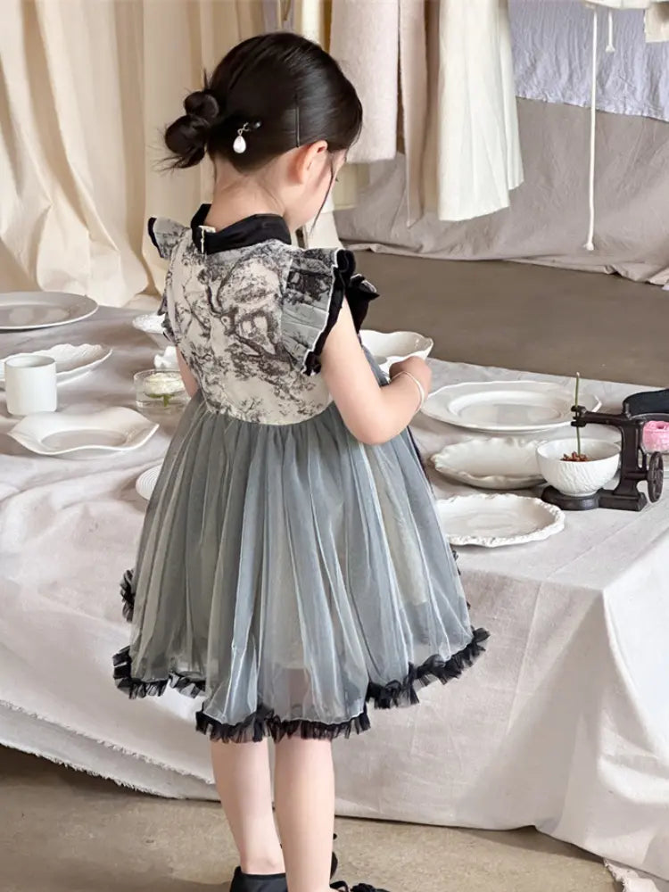 A young girl with her hair in a bun is shown from behind wearing a grey and black Girls Traditional Ink Painting Sleeveless Layered Tulle Qipao Dress  with a tulle skirt, standing next to a table set with white dishes.