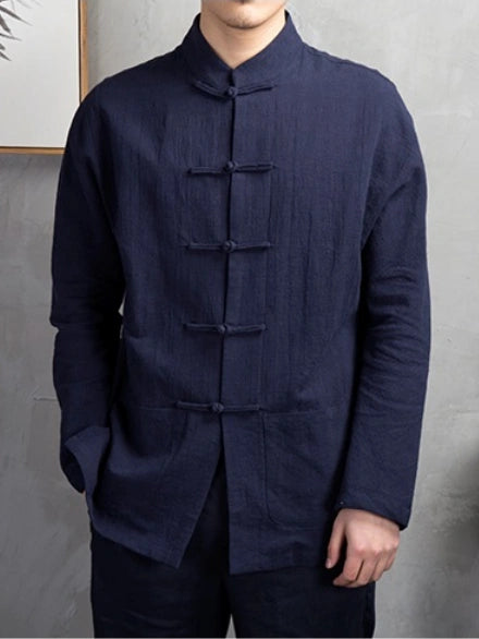 A man wearing a dark navy blue, long-sleeved traditional Chinese-style kongfu shirt with a mandarin collar and five front frog closures, paired with dark trousers, set against a textured gray wall.