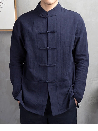 A man wearing a dark navy blue, long-sleeved linen tang kong fu shirt with traditional Chinese frog closures and a mandarin collar, standing against a textured gray wall.