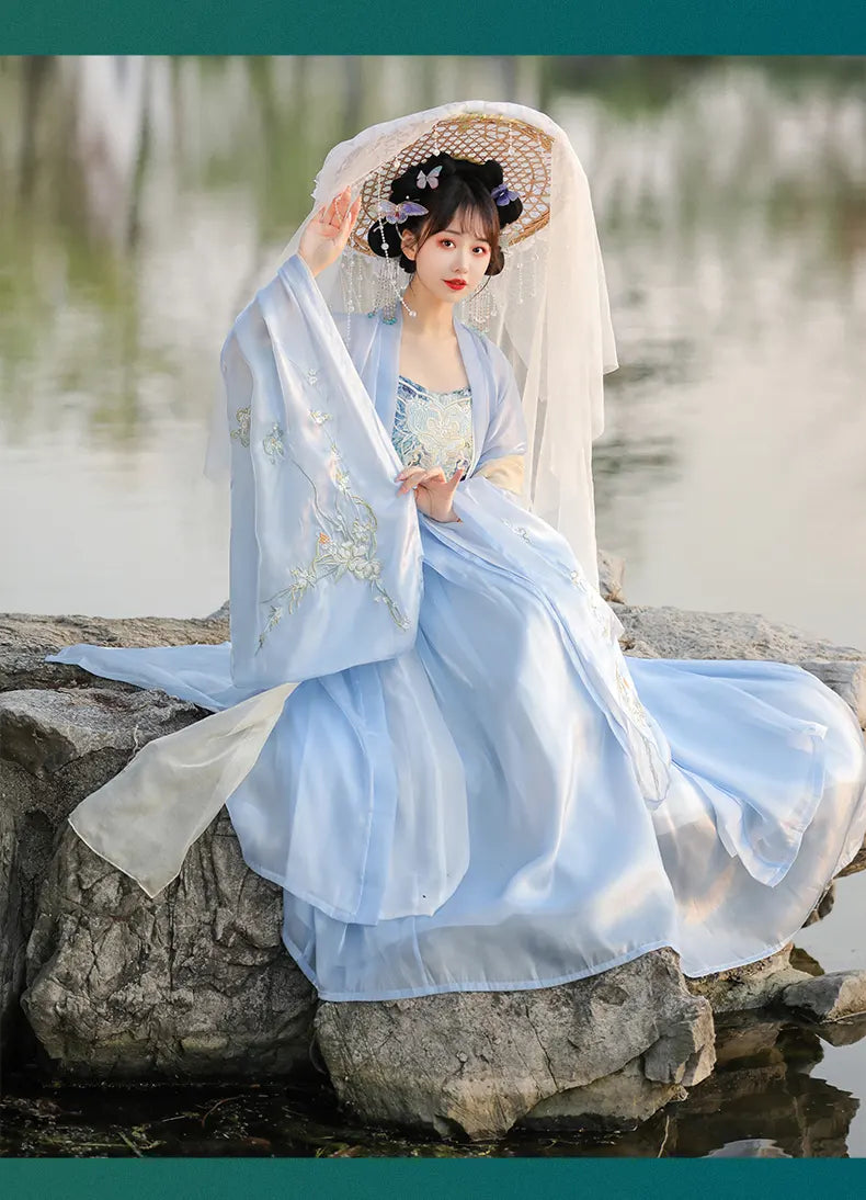 Woman in traditional light blue Chinese Tang Dynasty Ruqun Set Hanfu Dress with white veil sitting on a stone by a water body