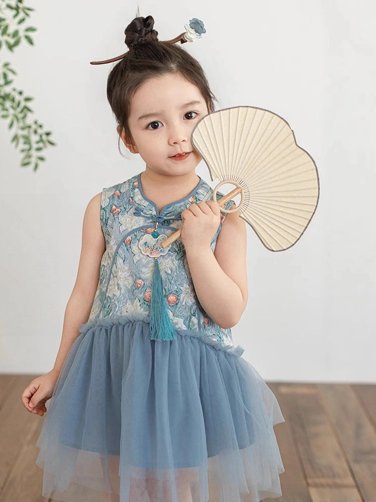 A young girl with her hair in a bun and a decorative hair stick is wearing a sleeveless light blue Girls Traditional Chinese Floral Sleeveless Tulle Qipao, holding a paper fan. She is standing in a room with a light-colored wall and wooden floor.