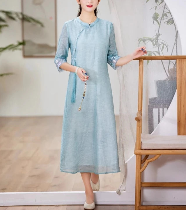 A chinese Woman wearing a light blue Loose A line Cheongsam Qipao Dress  standing indoors with a wooden chair and plant in the background.