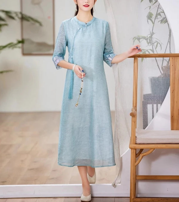 A chinese Woman wearing a light blue Loose A line Cheongsam Qipao Dress  standing indoors with a wooden chair and plant in the background.