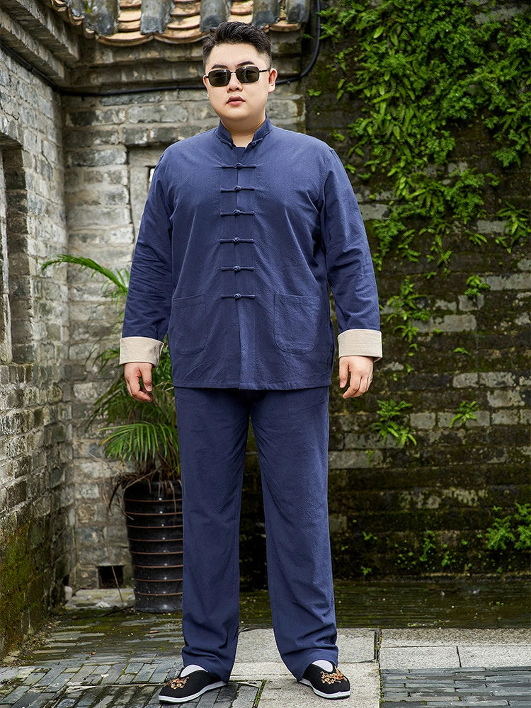 A man wearing a navy blue traditional Chinese-style two-piece set with beige cuffs, standing outdoors in front of a stone wall with green ivy.