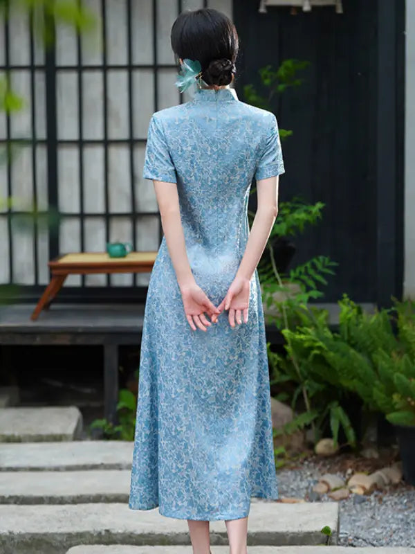 Woman wearing a light blue Modern Floral Short Sleeve A-Line Qipao Dress  standing outdoors with greenery and a building in the background.