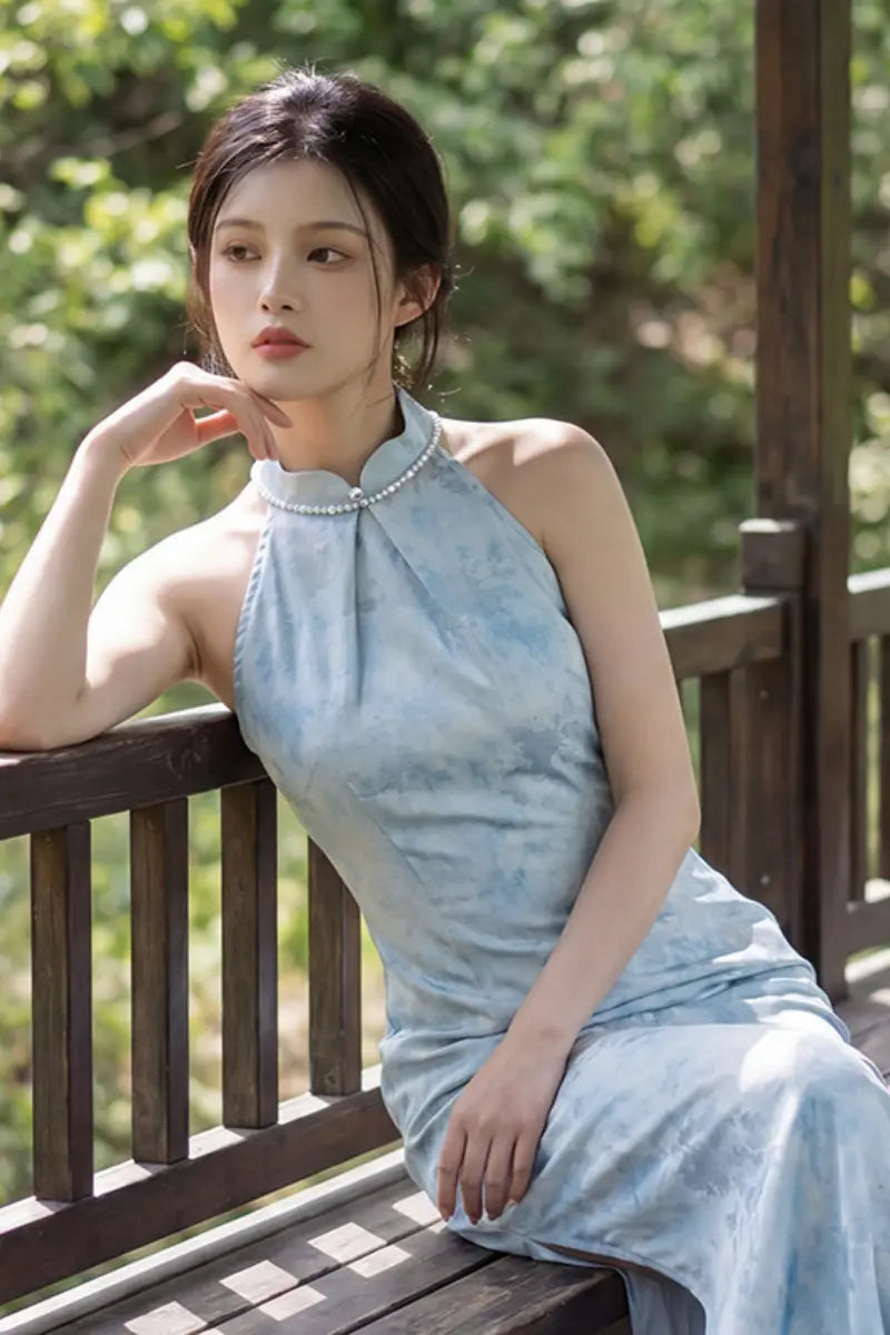 A woman wearing a light blue sleeveless high-neck Modern Pearl-Embellished Halter-Style Qipao  dress with a textured pattern, sitting on a wooden bench in an outdoor setting with green foliage in the background.