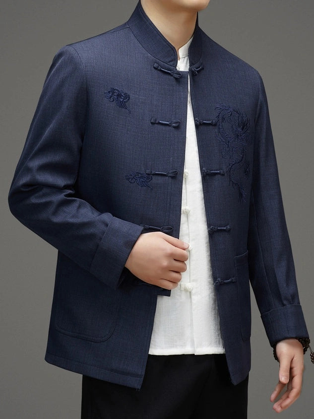 A man wearing a navy blue traditional Chinese jacket with embroidered details and frog closures, layered over a white shirt and black trousers, photographed against a neutral gray background.