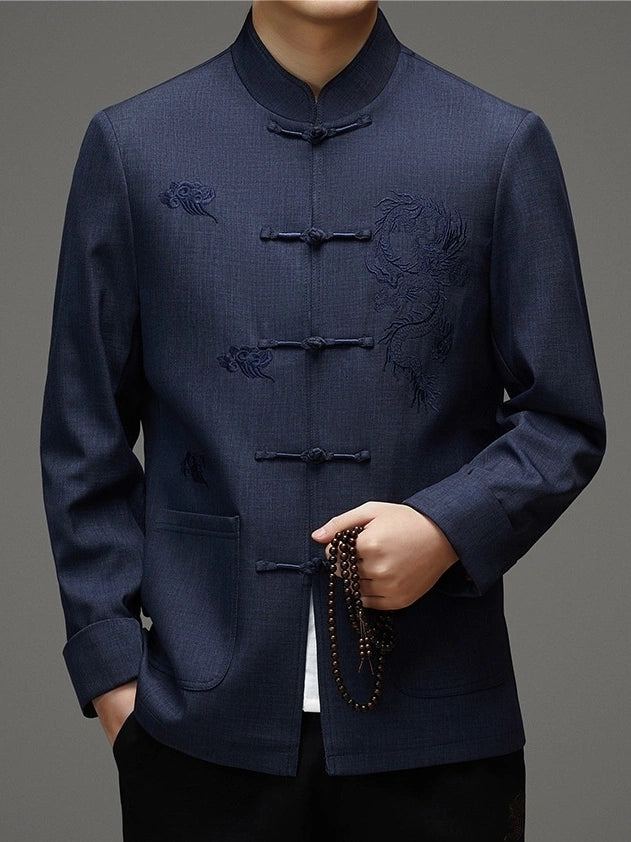 A man wearing a dark navy blue traditional Chinese jacket with dragon embroidery on the chest and dark wooden toggle closures, holding a beaded necklace against a neutral gray background.