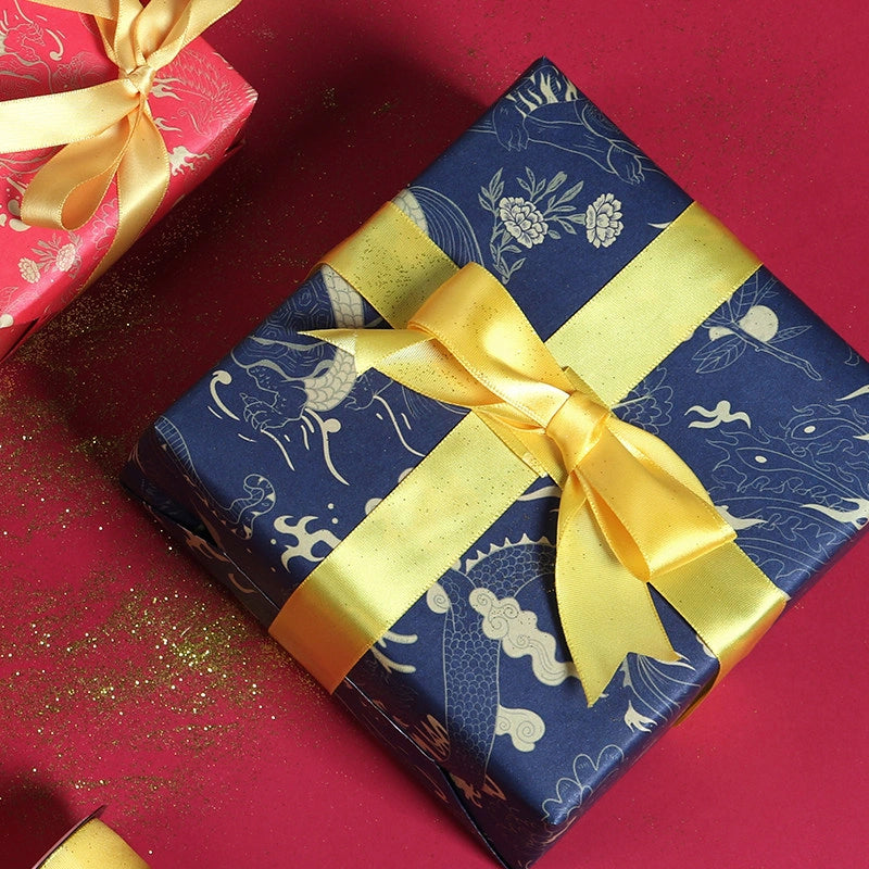 A square gift box wrapped in dark blue paper with a gold dragon Phoenix pattern and tied with a wide yellow satin ribbon, placed on a red surface with gold glitter. Part of another red gift box with a gold bow is visible in the top left corner.