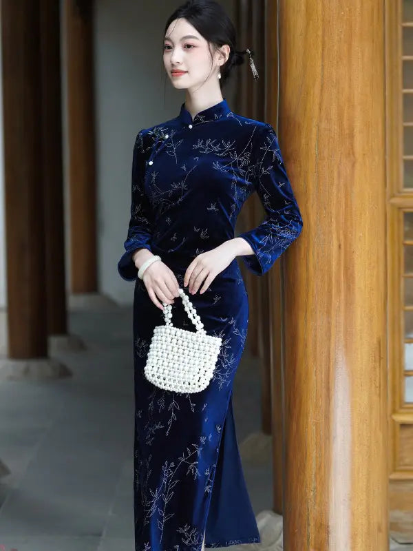 A woman wearing a dark blue Velvet Long Sleeve Qipao cheongsam dress with silver floral embroidery, holding a white woven handbag, standing in front of a wooden column.