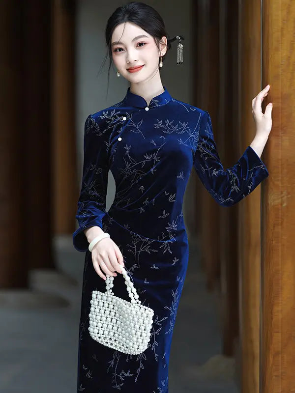 A woman models a dark blue Velvet Long Sleeve Qipao  cheongsam dress with light embroidery, holding a white woven handbag against a warm wooden background.