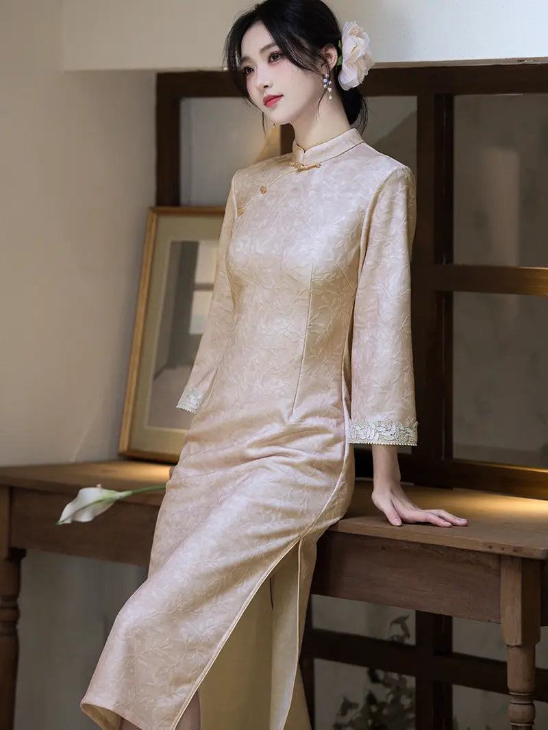 A woman wearing a light beige satin cheongsam dress with long sleeves and a high collar, leaning on a wooden table in an indoor setting.