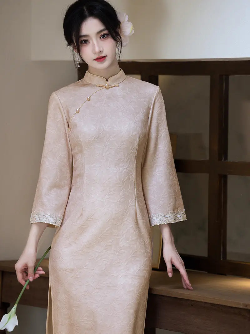 A woman wearing a light beige textured cheongsam dress with three-quarter sleeves and lace trim at the cuffs, standing in a warm-toned indoor setting.