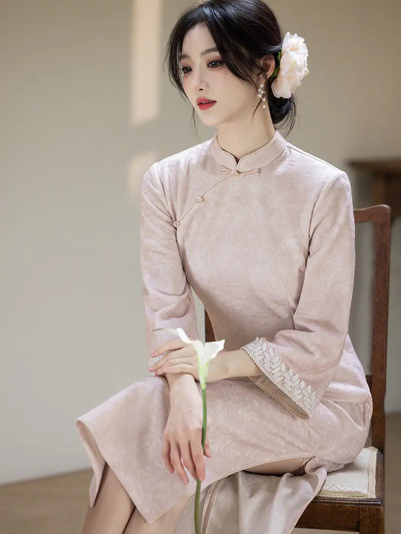 A woman seated on a wooden chair wearing a pale blush pink traditional Chinese cheongsam dress with lace trim on the cuffs and a white flower in her hair, holding a white flower stem.