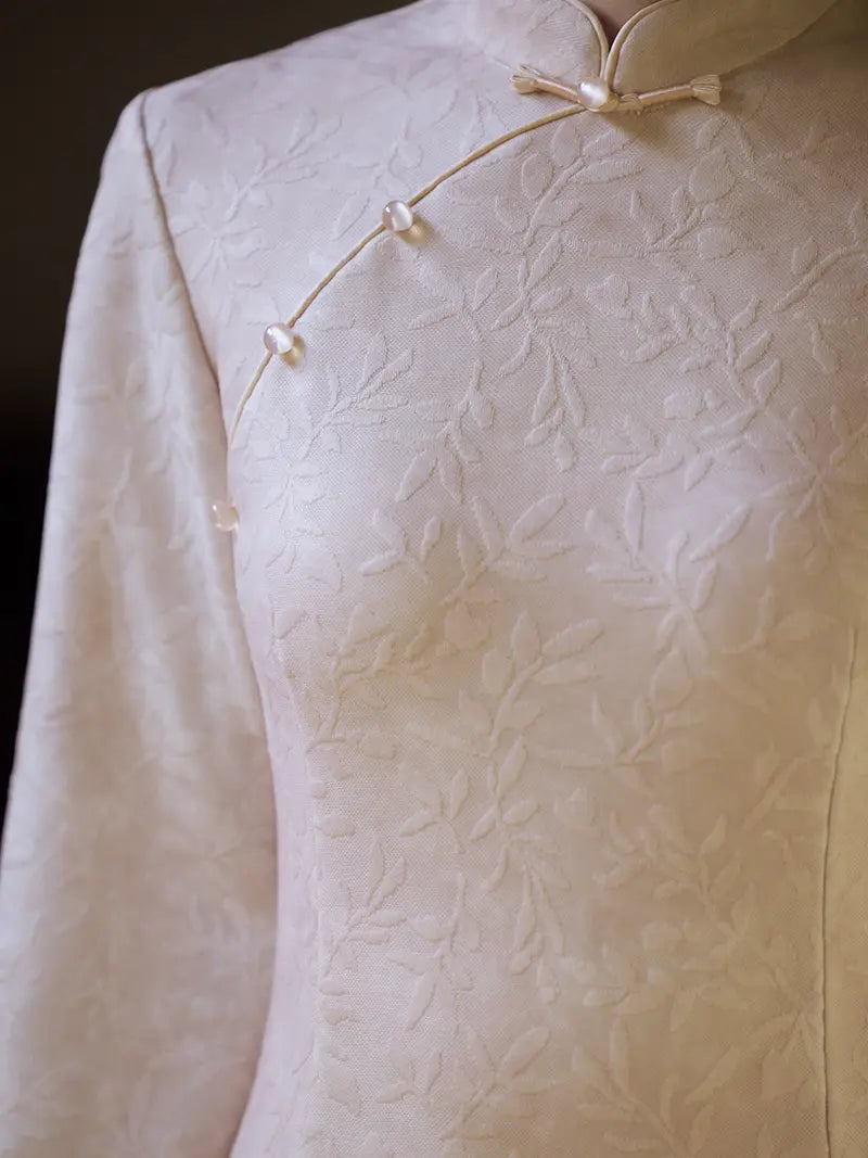 Close-up of a cream-colored traditional Chinese cheongsam dress with a subtle floral jacquard pattern and decorative buttons along the collar.