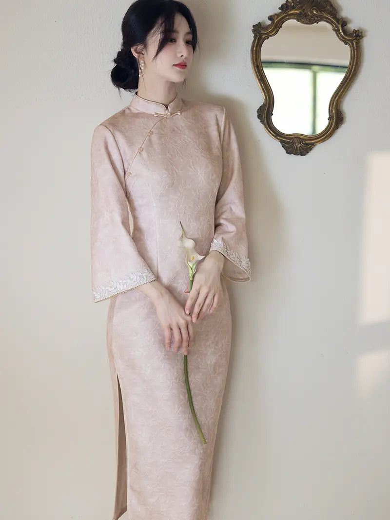 A woman wearing a light blush pink satin cheongsam (qipao) dress with lace trim on the cuffs, standing against a cream-colored wall next to an ornate gold mirror.