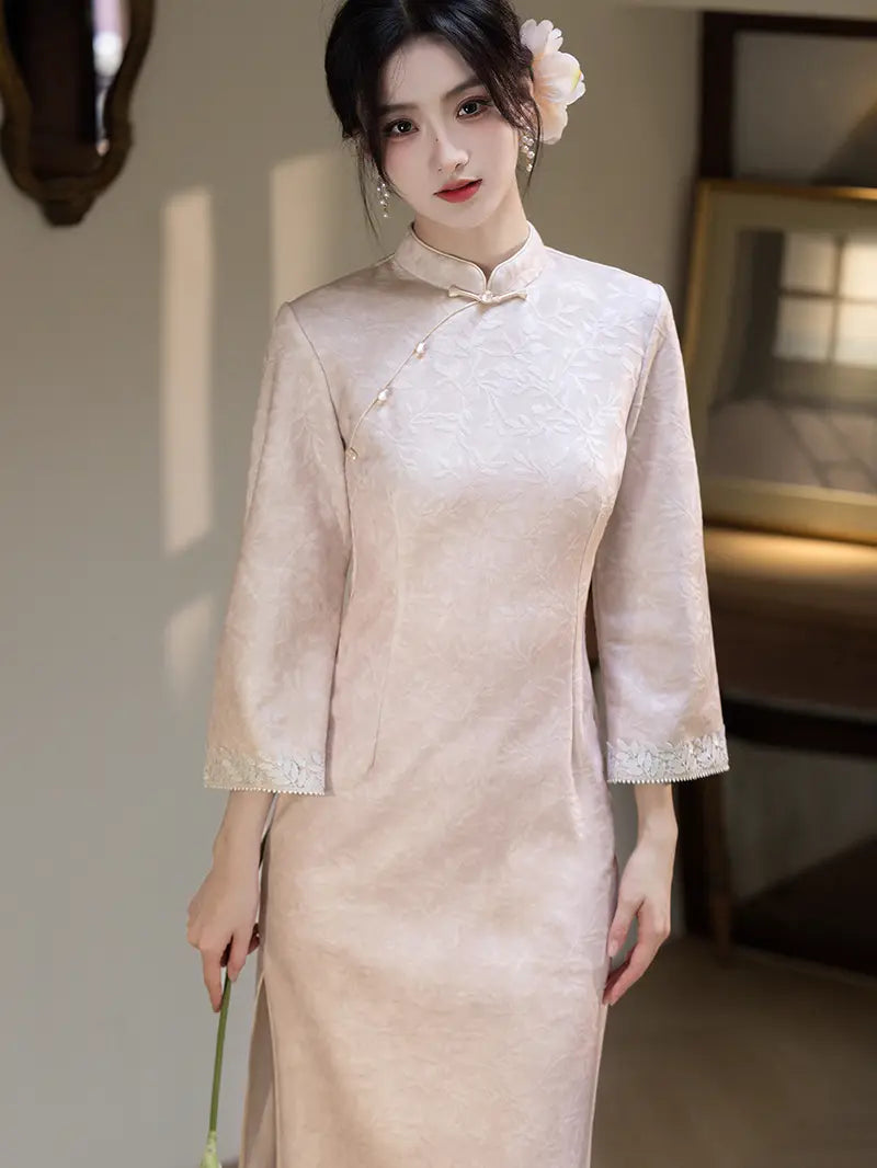 A woman wearing a pale blush pink textured Chinese cheongsam (qipao) dress with three-quarter sleeves and a high collar, standing in a softly lit indoor setting.