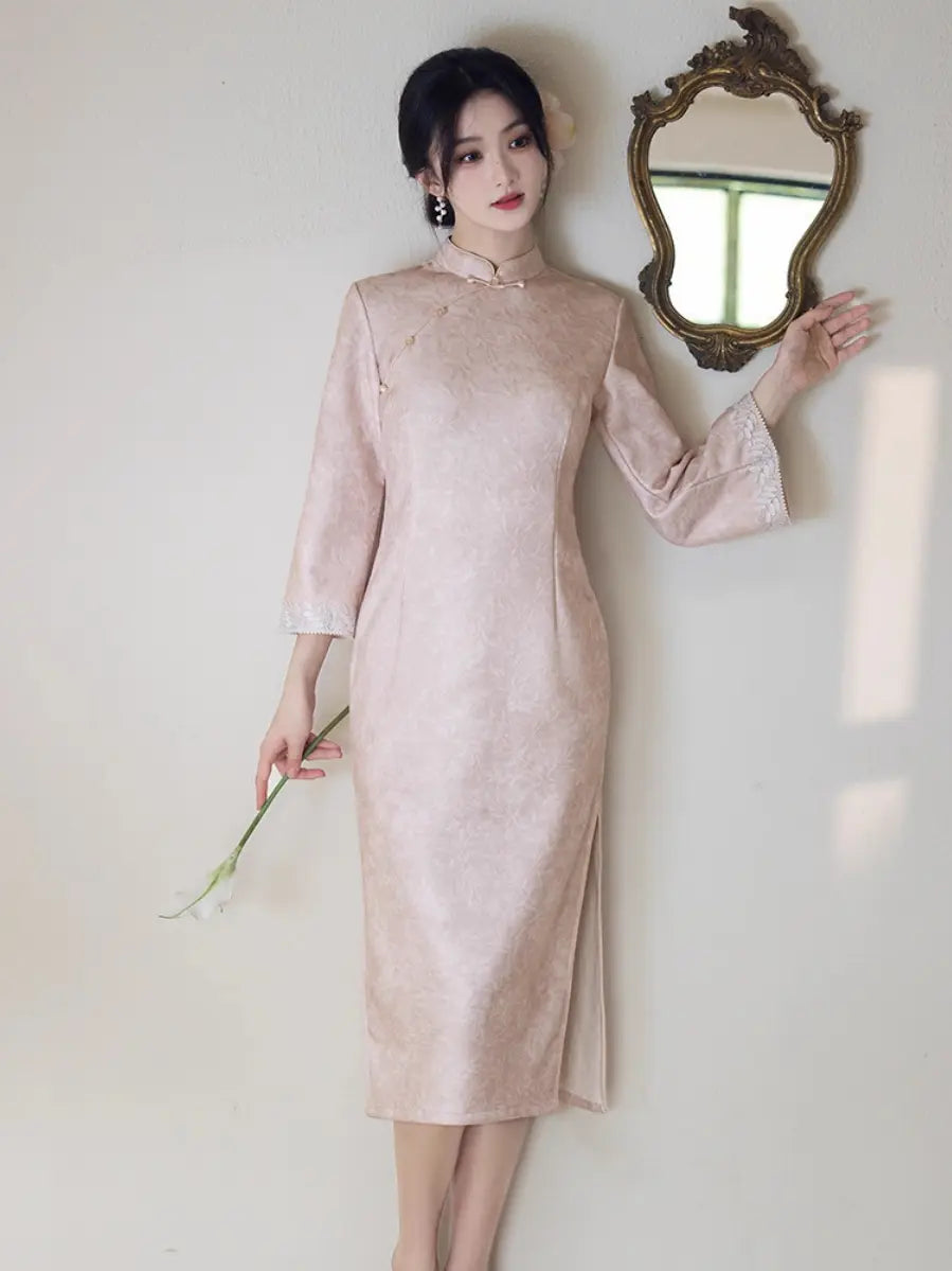 A woman wearing a light pink traditional Chinese cheongsam dress stands against a plain wall, holding a small white flower and touching an ornate gold mirror.
