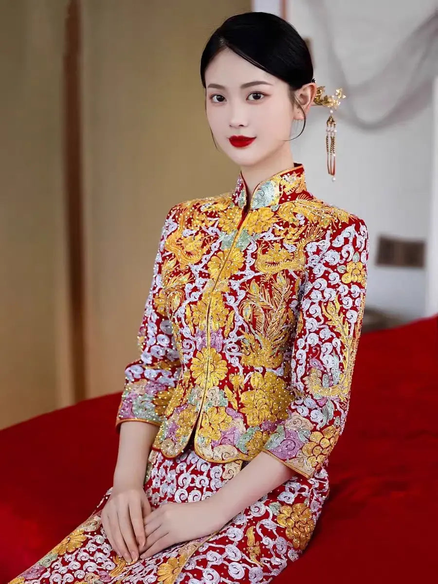 A woman seated on a red bench, wearing a traditional Chinese Qun Kwa dress with gold and silver thread embroidery featuring dragon and phoenix motifs.