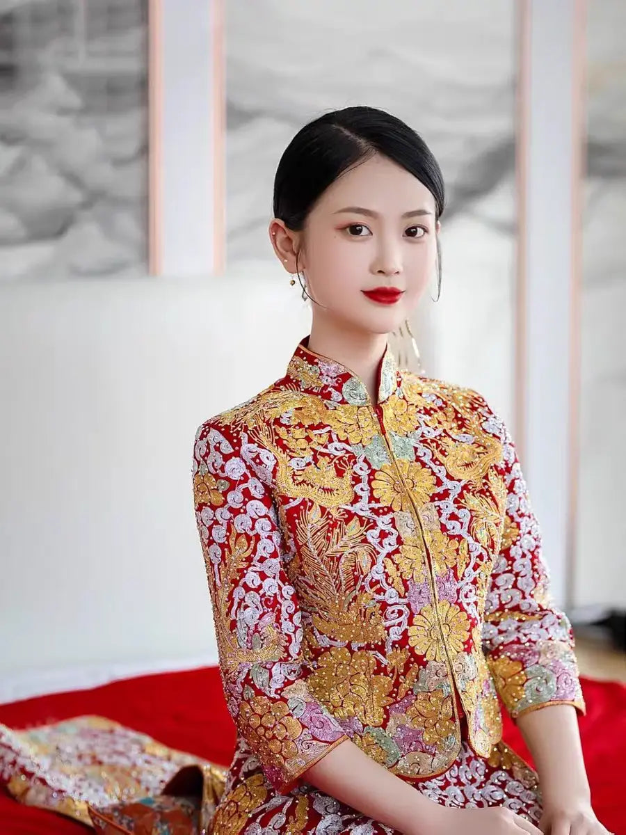 A woman seated on a red bench, wearing a traditional Chinese Qun Kwa dress with gold and silver thread embroidery featuring dragon and phoenix motifs.