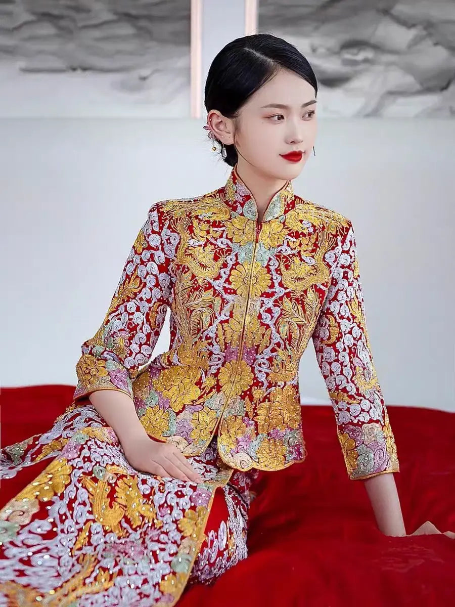 A woman seated on a red bench, wearing a Multicolor Embroidered Chinese Bridal Outfit  with gold and silver thread embroidery featuring dragon and phoenix motifs.