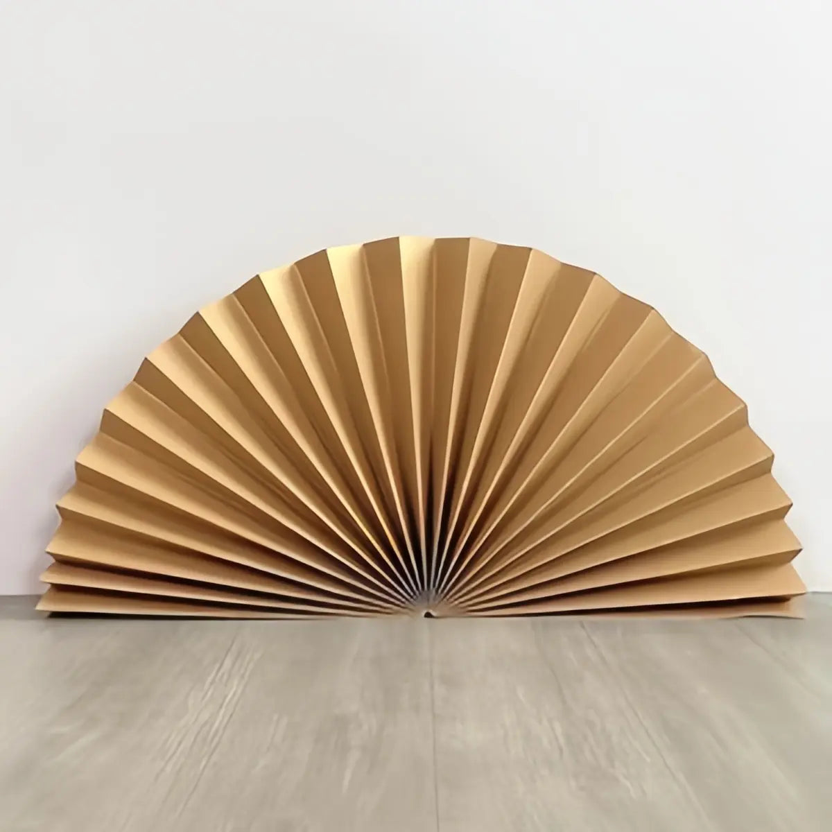 A gold-colored pleated accordion wall fan, shown open and standing on a light gray wooden floor against a white wall.