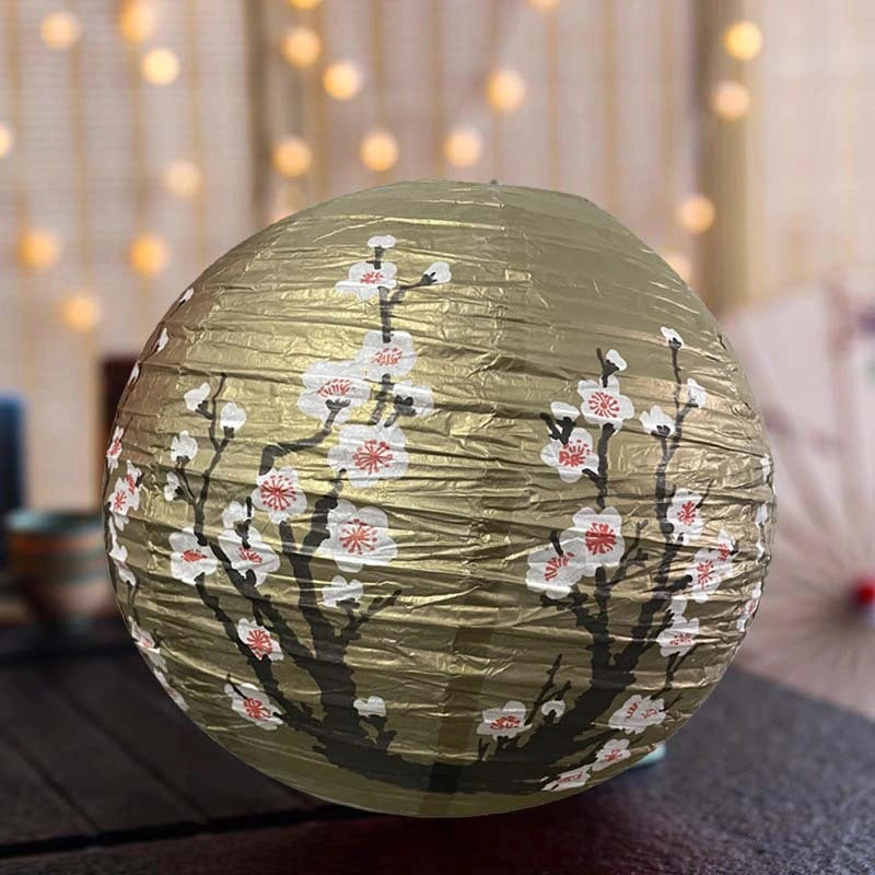 A round gold paper lantern decorated with black branches and white cherry blossoms with pink centers, shown in a softly blurred indoor setting with warm string lights in the background.