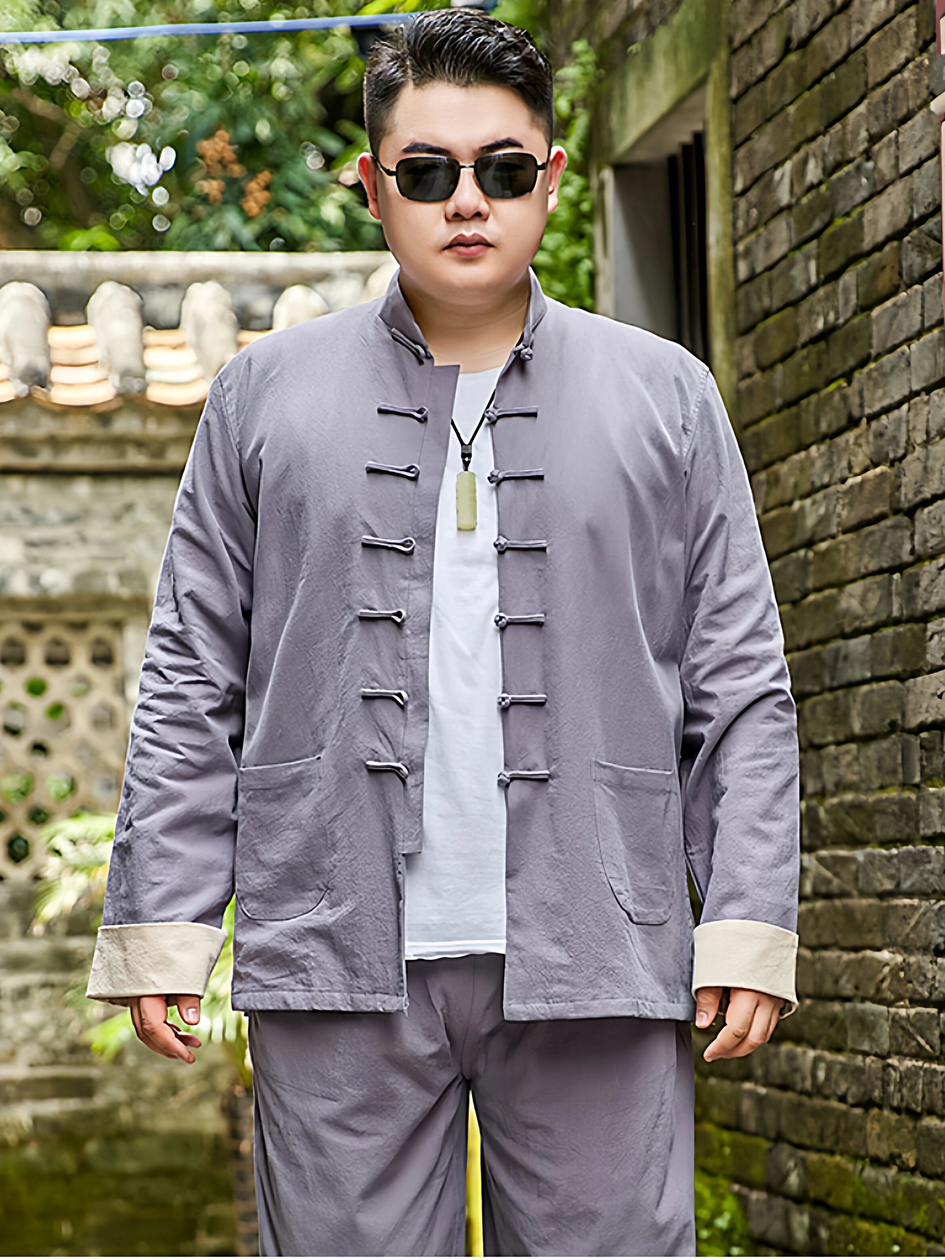 A man wearing a plus size light gray traditional Chinese-style jacket and matching pants over a white t-shirt, standing outdoors in front of a brick wall.
