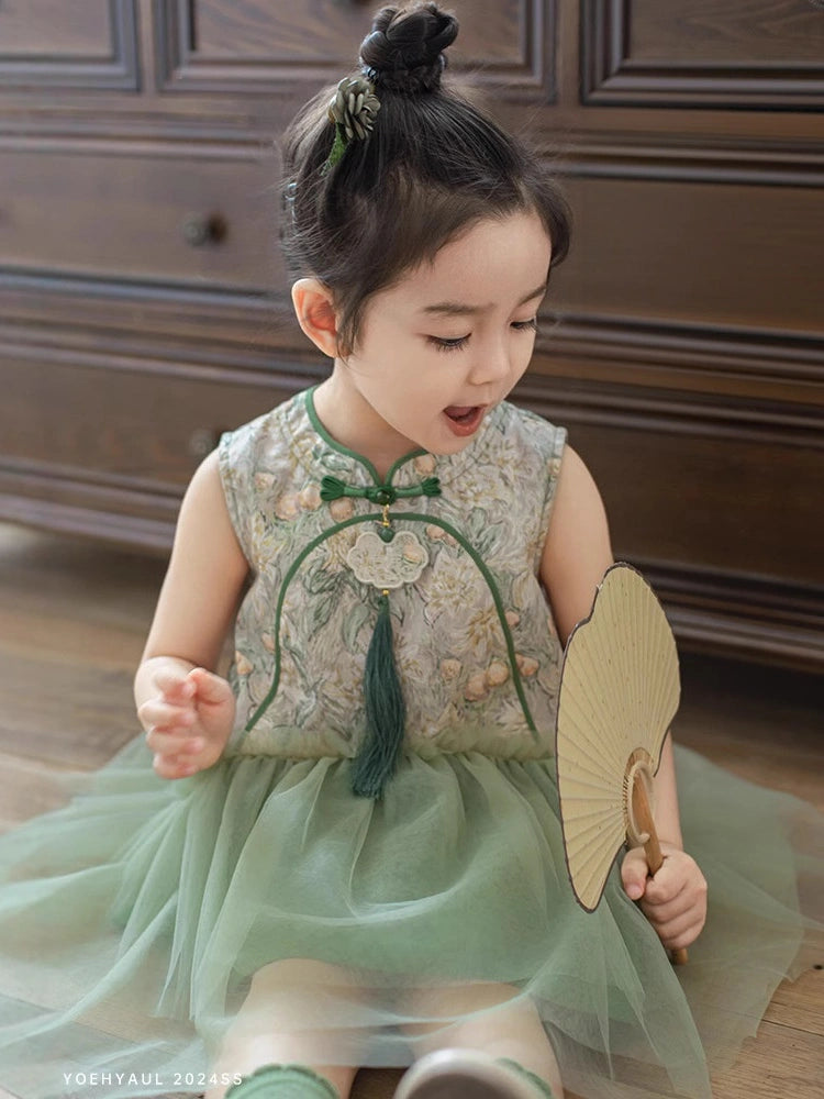 A toddler girl wearing a sleeveless light green Girls Traditional Chinese Floral Sleeveless Tulle Qipao  dress with floral embroidery and a tulle skirt, sitting on a wooden floor and holding a folding fan.
