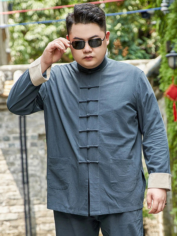 A man wearing a dark slate blue-gray plus size traditional Chinese mandarin collar jacket with black frog closures and beige turned-back cuffs, paired with matching pants and sunglasses, standing outdoors against a blurred stone wall and greenery background.